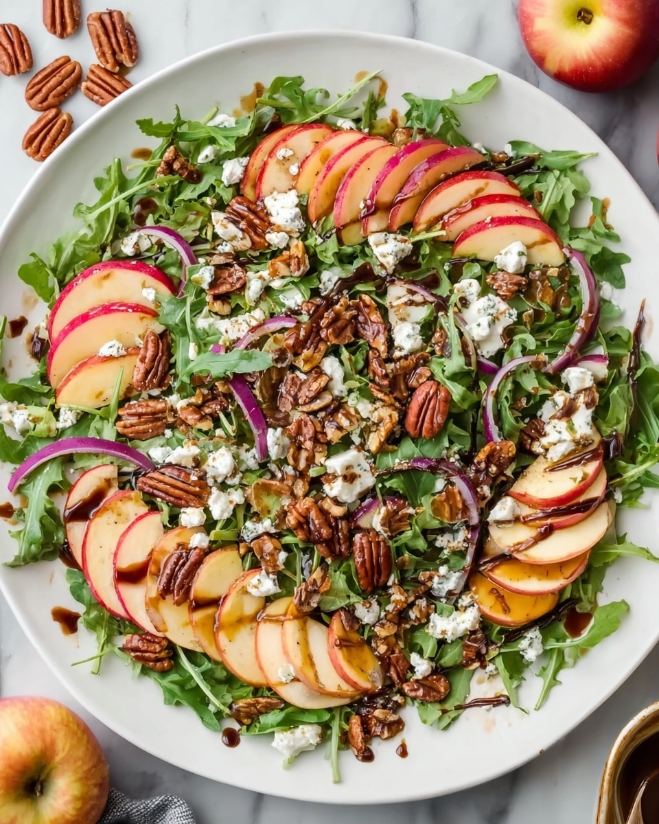 A white plate holds a fresh salad with multiple layers starting with a bed of green leafy arugula. On top of this are thin slices of red and yellow apple, arranged in curved rows around the plate’s edge. Scattered pecans add a rich brown color and rough texture across the salad. Small chunks of white goat cheese are spread unevenly over the top, bringing a soft, crumbly element. Thin strips of purple-red onion add a splash of color among the nuts and cheese. The salad is lightly drizzled with a glossy, dark balsamic dressing that pools in some areas. The plate sits on a white marbled surface with some fresh apples and pecans visible nearby. photo taken with an iphone --ar 4:5 --v 7
