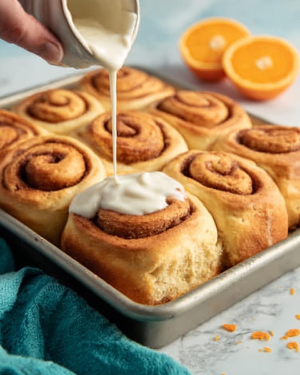 The image shows a white baking tray with six golden brown cinnamon rolls inside. The rolls have a soft, fluffy texture and a visible spiral pattern on top. A woman's hand is pouring white cream cheese glaze from above onto the center cinnamon roll, the glaze looking smooth and thick as it drips down. In the background, there are halved oranges on a white marbled surface. photo taken with an iphone --ar 4:5 --v 7