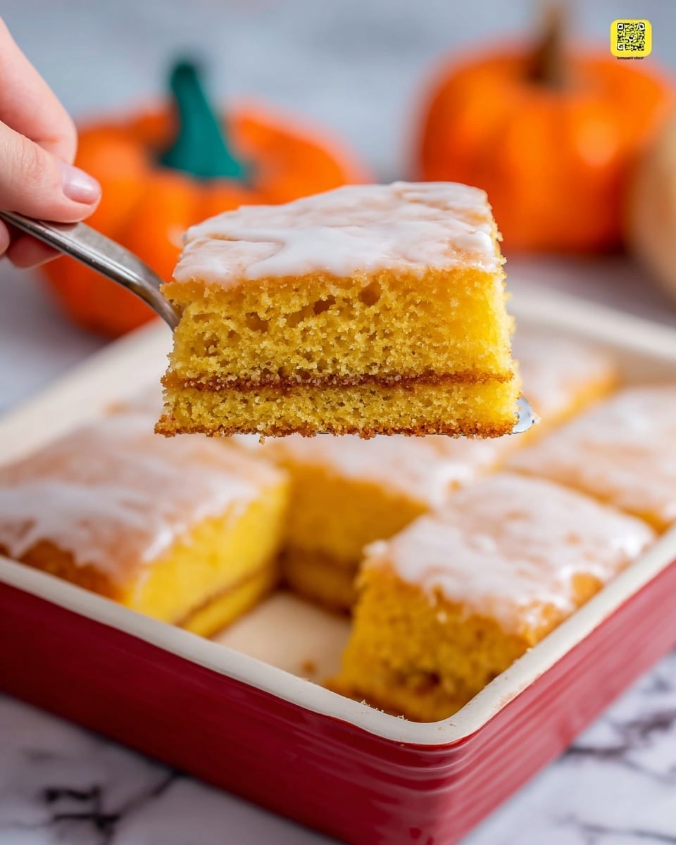 A close-up of a square piece of yellow cake being lifted by a woman’s hand using a spatula. The cake has two thick layers of moist, soft yellow sponge with a thin layer of brown filling in the middle. The top layer is covered with a smooth, light, white glaze that slightly drips down the sides. The cake piece is held above a white baking dish with a red rim, showing the same layered cake cut into squares inside the dish. In the background, bright orange small pumpkins sit on a white marbled surface. photo taken with an iphone --ar 4:5 --v 7