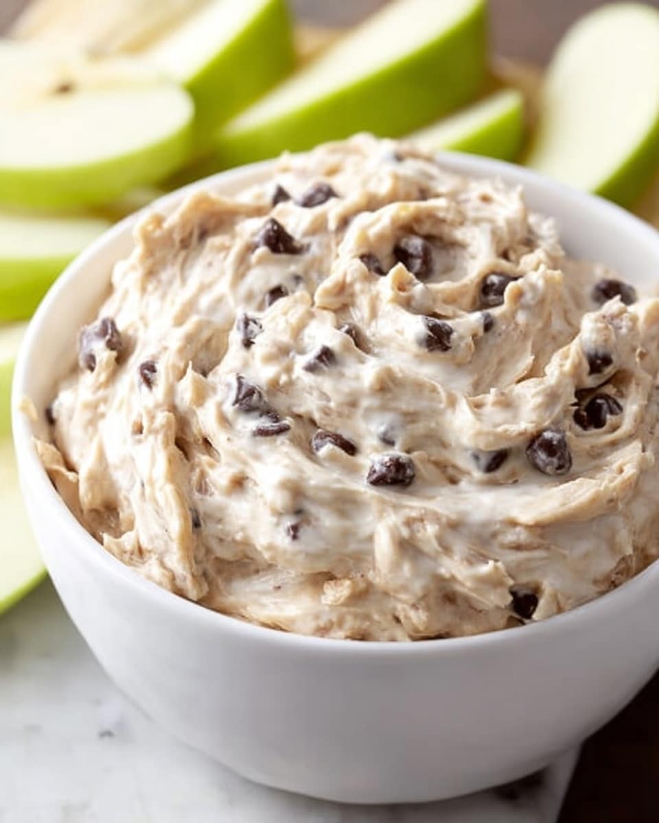 A close-up of a white bowl filled with creamy, light beige spread mixed with small bits of dark raisins and pieces of walnuts. The spread has a smooth but slightly textured surface with swirls and folds showing its thick, soft consistency. Around the bowl, there are slices of green apples placed upright, adding a fresh color contrast against the spread. The bowl sits on a white marbled surface. Photo taken with an iphone --ar 4:5 --v 7