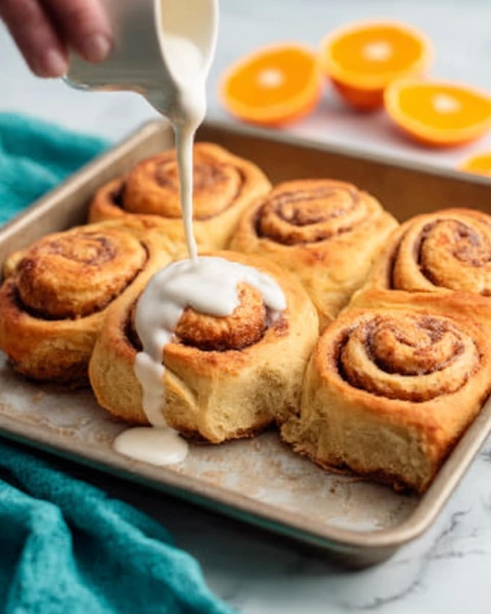 A close-up image showing six golden brown cinnamon rolls in a light-colored metal baking tray. One cinnamon roll at the front center is being drizzled with thick, white cream cheese icing from above, held by a woman's hand. The rolls have a soft, fluffy texture with a visible swirl pattern of cinnamon inside. In the background, there are halves of bright orange fruit placed on a white marbled surface. A teal cloth is partially visible at the bottom left corner. Photo taken with an iphone --ar 4:5 --v 7