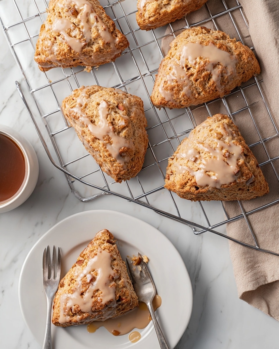 The image shows five golden brown scones with a rough, crumbly texture topped with a light brown glaze that looks creamy and slightly glossy. Four whole scones rest on a metal cooling rack, spaced apart to show their triangular shapes with bits of white inside, likely pieces of fruit or nuts. One scone sits on a white plate in the lower right, broken into two parts with the glaze dripping slightly, and a silver fork rests on the plate next to the scone pieces. The scene is set on a white marbled surface, and a folded light brown cloth is partially visible in the upper right corner. Photo taken with an iphone --ar 4:5 --v 7