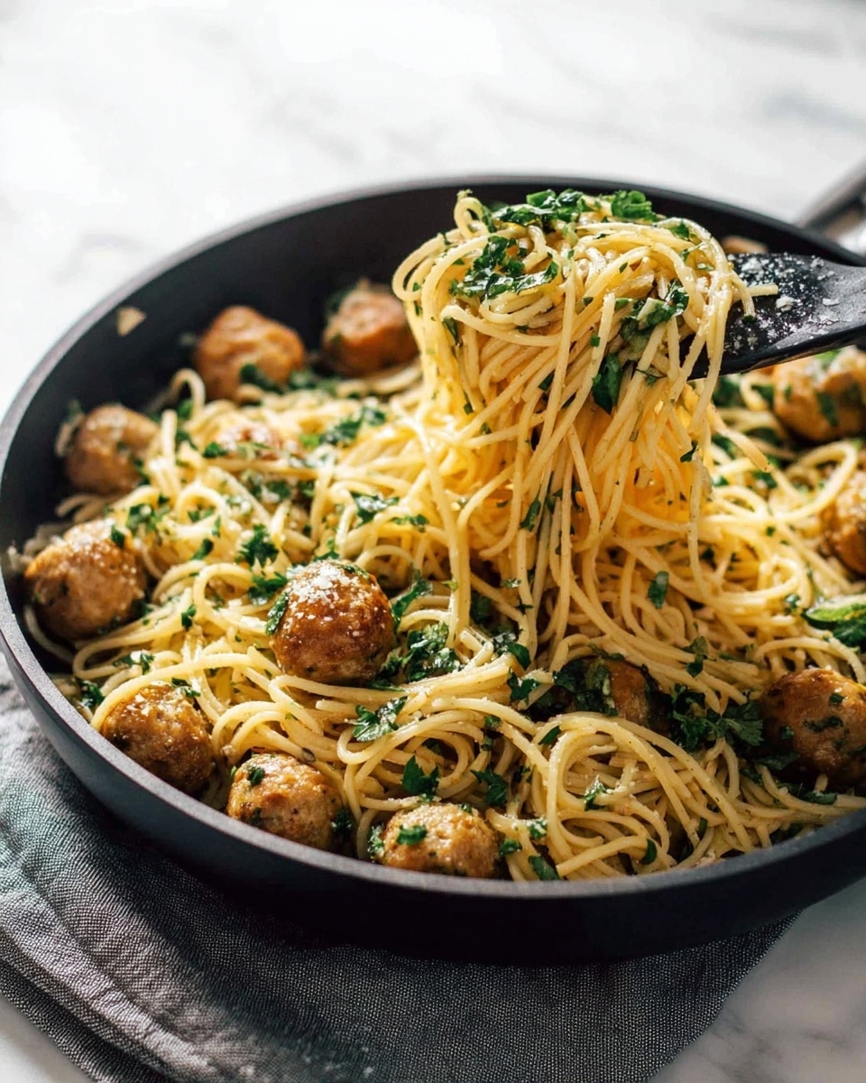 The image shows a black pan filled with spaghetti pasta mixed with small round golden-brown meatballs and fresh green chopped herbs scattered evenly throughout. The noodles are a light yellow color and look glossy, coated with a sauce that makes them appear slightly shiny. Some spaghetti and herbs are being lifted out of the pan with a black utensil, showing the texture and length of the noodles. The pan sits on a white marbled surface with a gray cloth napkin nearby. photo taken with an iphone --ar 4:5 --v 7