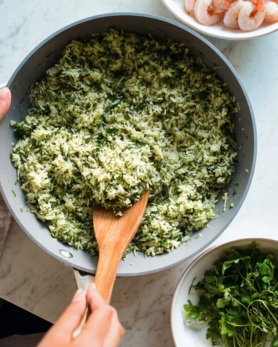 A close-up of cooked green herb rice inside a gray pan, with the rice showing a mix of light green and white grains blended with small dark green herb pieces spread evenly in one thick layer filling the pan; a wooden spatula is stirring the rice in the lower right part of the pan, held by a woman's hand; to the right, a white bowl with fresh green herbs and pink shrimp is partly visible; the setting is on a white marbled surface; photo taken with an iphone --ar 4:5 --v 7