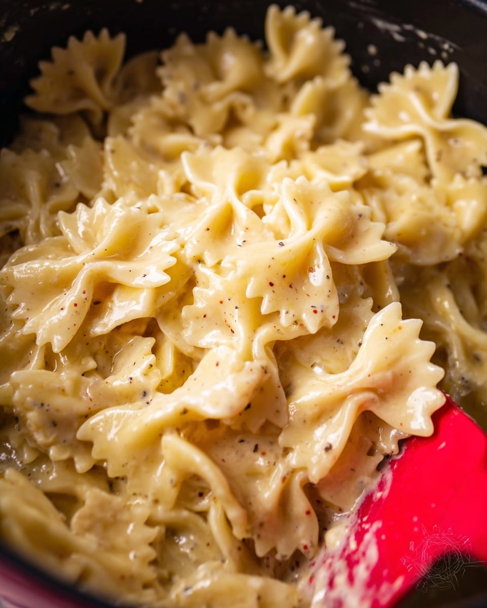 The image shows a close-up of creamy farfalle pasta in a pot, with one visible layer of bow-tie shaped pasta covered in smooth yellow cheese sauce with tiny specks of pepper. The pasta appears soft and glossy, and a red spatula is partially visible on the right side, mixing or scooping the dish. The background is a soft focused black inside of the pot. The photo taken with an iphone --ar 4:5 --v 7