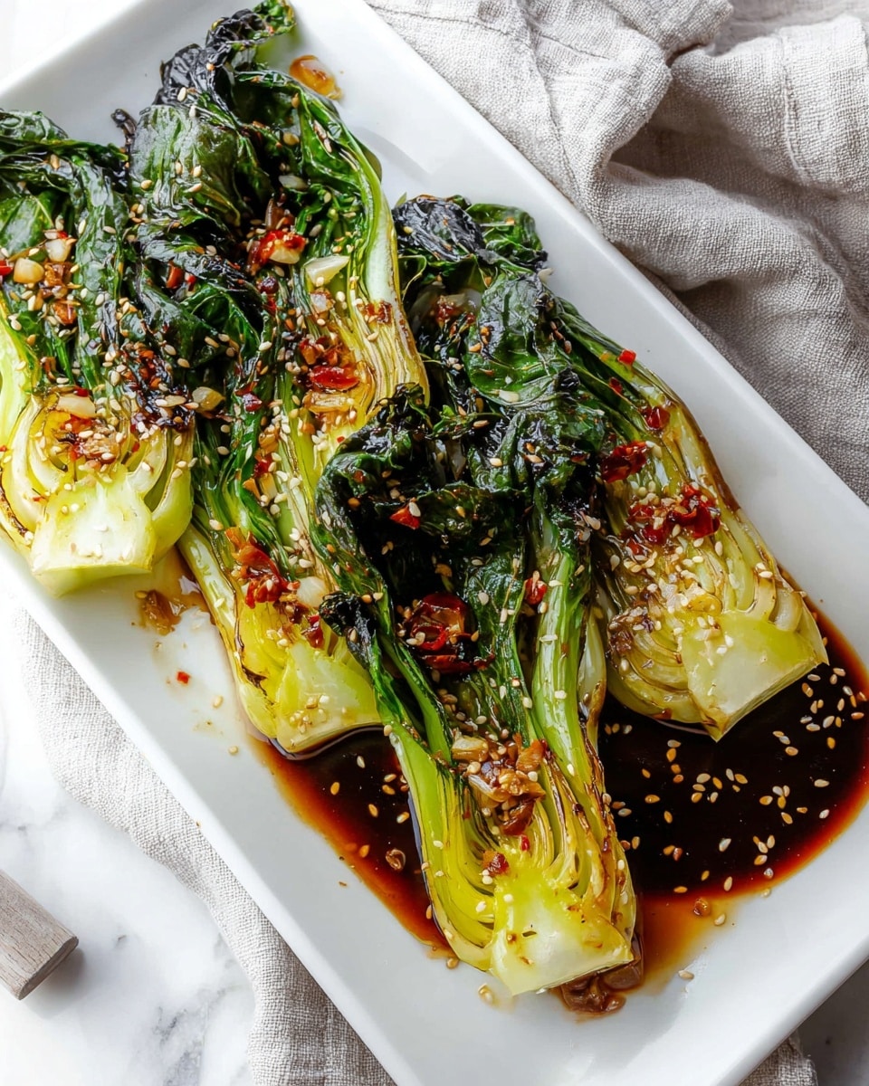 A white rectangular plate holds four pieces of baby bok choy, each cut in half lengthwise and arranged side by side. The bok choy leaves are dark green with a wilted texture, while the stalks are light green to pale yellow with a slightly charred or caramelized surface. A glossy dark brown sauce pools around the base of the bok choy, with small chunks of red chili flakes and garlic scattered on top. Light tan sesame seeds are sprinkled over the entire dish, adding a touch of texture. The plate sits on a white marbled surface with a soft, light gray cloth draped in the background. Photo taken with an iphone --ar 4:5 --v 7