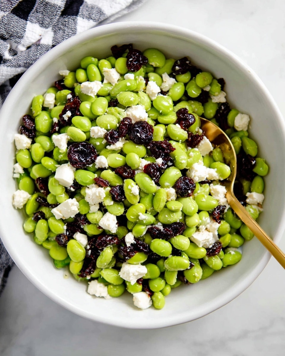 A white bowl filled with a salad made of bright green edamame beans, scattered with small chunks of white cheese and dark dried cherries. The edamame beans create the base layer, with the white cheese and cherries randomly mixed throughout. A gold spoon is placed in the bowl, slightly buried in the salad, with a black and white checkered cloth in the background on a white marbled surface. photo taken with an iphone --ar 4:5 --v 7