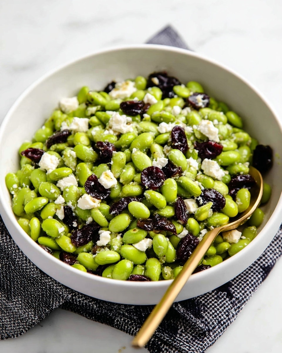 A white bowl filled with a bright green edamame salad mixed with small chunks of white feta cheese and dark purple dried cherries scattered evenly throughout. The salad appears fresh and lightly coated with a hint of oil, creating a slight shine on the edamame beans. A gold spoon is placed inside the bowl on the right side, partially inserted into the salad. The bowl sits on a black and white checkered cloth, all set against a white marbled surface. photo taken with an iphone --ar 4:5 --v 7