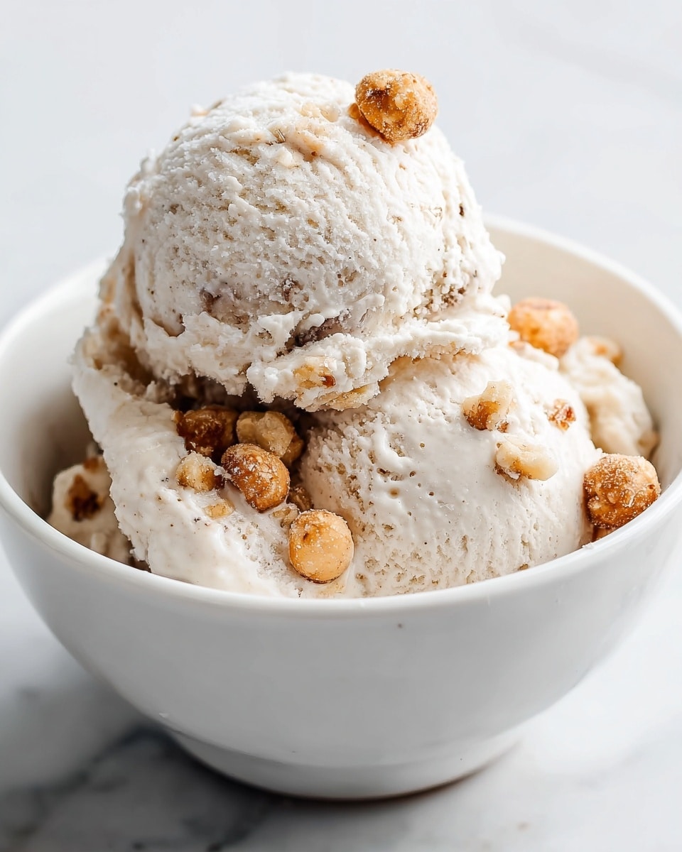 The image shows a white bowl filled with two scoops of creamy, light beige ice cream that has a slightly crumbly texture and some brown nut pieces mixed inside. On top of the scoops, there are several small, round, golden-brown nuts adding extra texture and color contrast. The bowl sits on a white marbled surface with a clean, bright background. Photo taken with an iphone --ar 4:5 --v 7