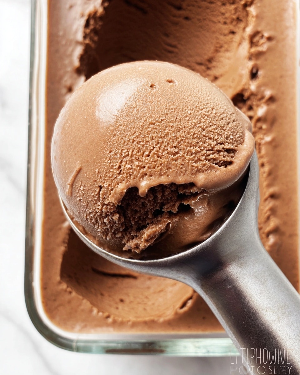 A close-up view of a single scoop of smooth, creamy chocolate ice cream being lifted by a shiny metal ice cream scooper from a rectangular clear glass container filled with the same chocolate ice cream, showing a rich, medium brown color with a slightly glossy texture and tiny ice crystals on the surface; the background surface is a white marbled texture. photo taken with an iphone --ar 4:5 --v 7