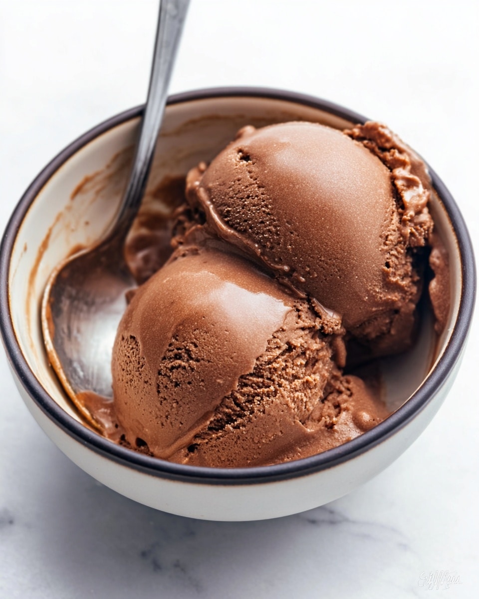 The image shows two scoops of smooth chocolate ice cream in a white bowl with a dark rim. The scoops are rich brown in color with a creamy and slightly melted texture around the edges. A shiny metal spoon is placed inside the bowl on the left side, with some melting ice cream visible on the bowl’s inner wall. The background is a white marbled surface. photo taken with an iphone --ar 4:5 --v 7